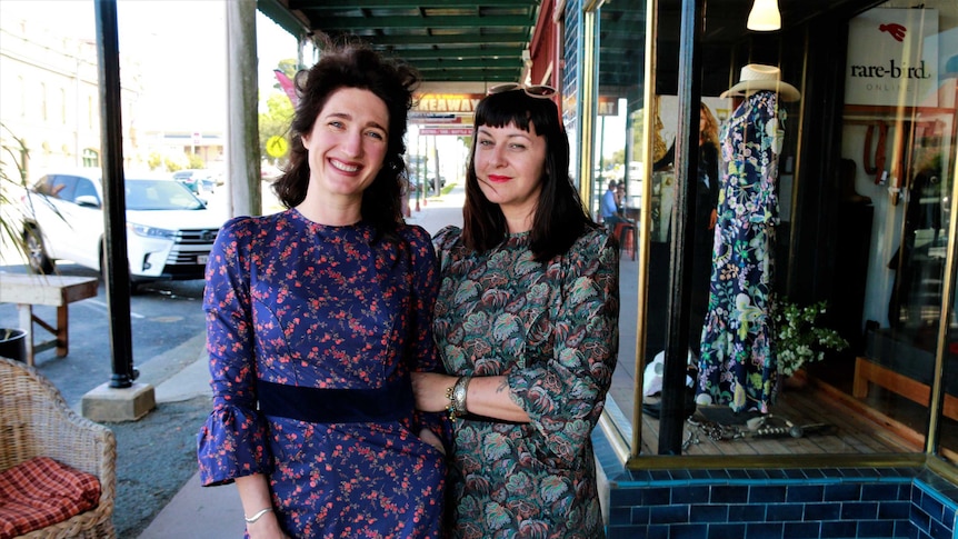 Two dark-haired women stand smiling while wearing colourful floral dresses outside their clothes shop in Braidwood, NSW.