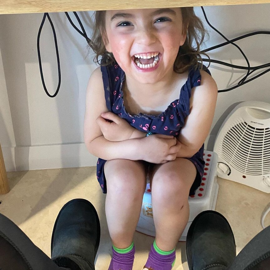 A little girl smiles up from under a computer table