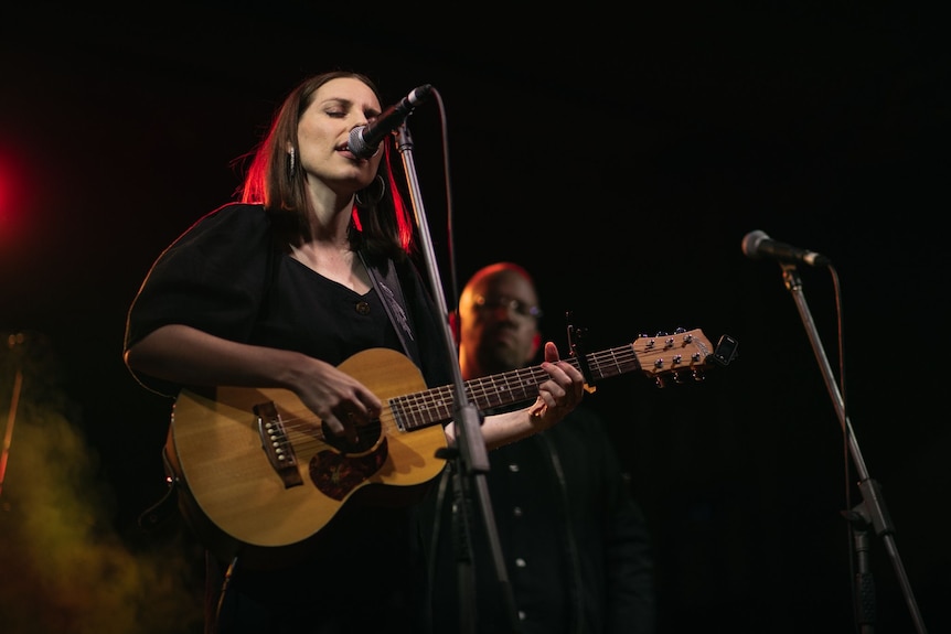 A woman stands at a microphone with an acoustic guitar singing. 