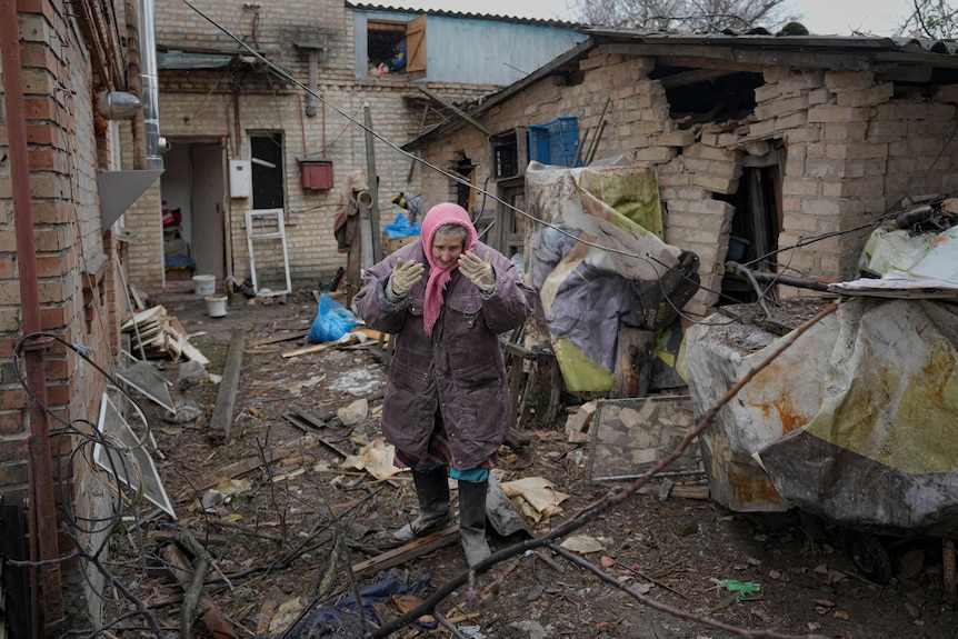 An elderly woman wearing winter clothes, boots and gloves stands among a destroyed building, bringing her hands to her eyes.