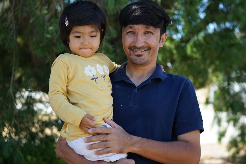 Ali Rahmani holds his toddler daughter in front of trees smiling