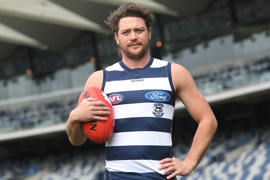 Jack Steven stands in an empty stadium holding a red football and wearing a Geelong uniform.