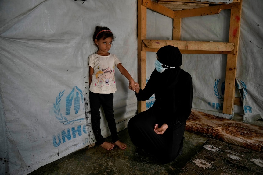 A woman kneels beside a small child leaning against the side of an aid tent. 