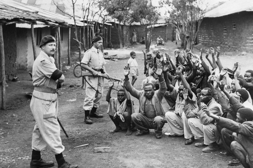B&W photo of two men in colonial uniforms with guns guarding about 20 African men, crouching with their hands up