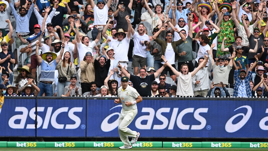 A cricket player in whites celebrates with crowd raising arms and cheering behind.