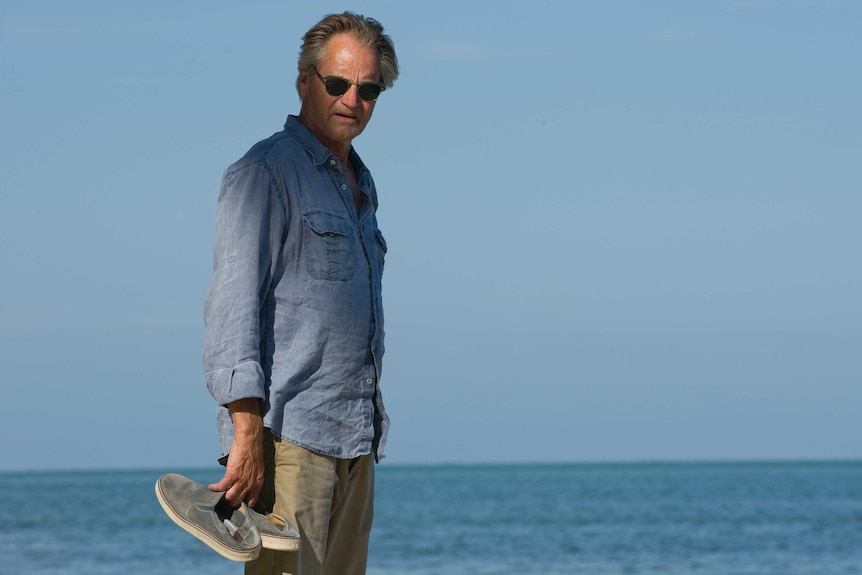 Sam Shepard holds shoes in his hand as he stands on a beach.