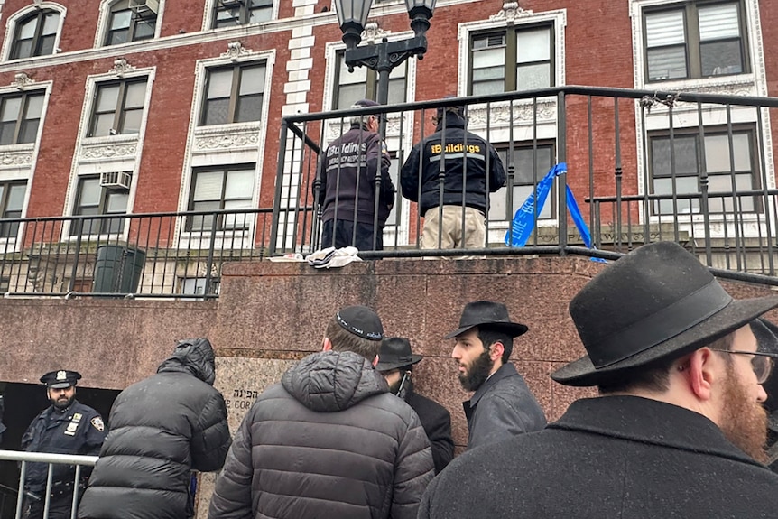 A group of Jewish men huddle outside near barricades outside a building. Police and inspectors on other side of barricades