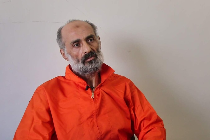 Older man with grey and black beard sits wearing orange prison uniform in front of dull cream wall.