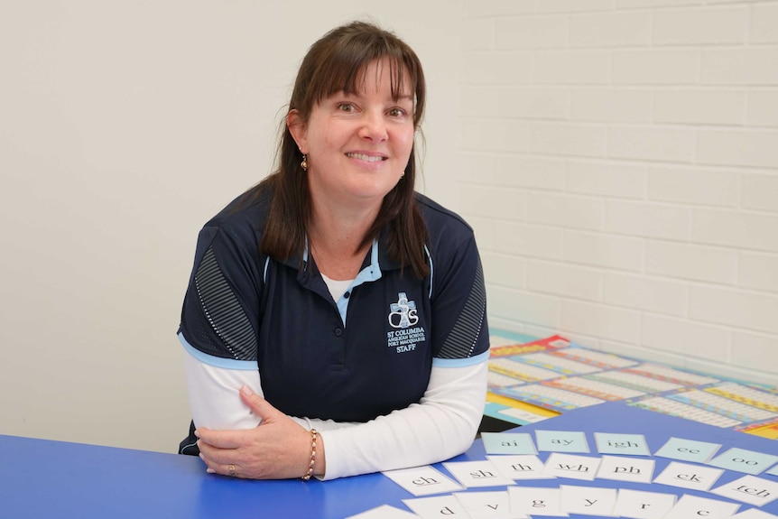 A female teacher sits smiling at a desk in a classroom, with letters on cards on the desk.