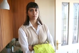 A mid-shot of a woman with a serious expression on her face and holding a hi-vis vest standing indoors.