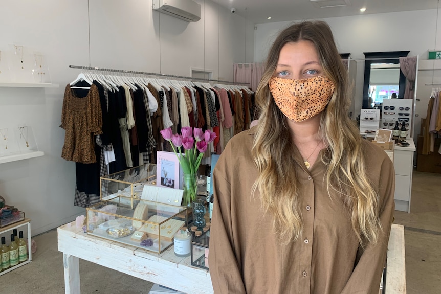 A woman with long hair in a shop featuring racks of clothes, wearing a mask.