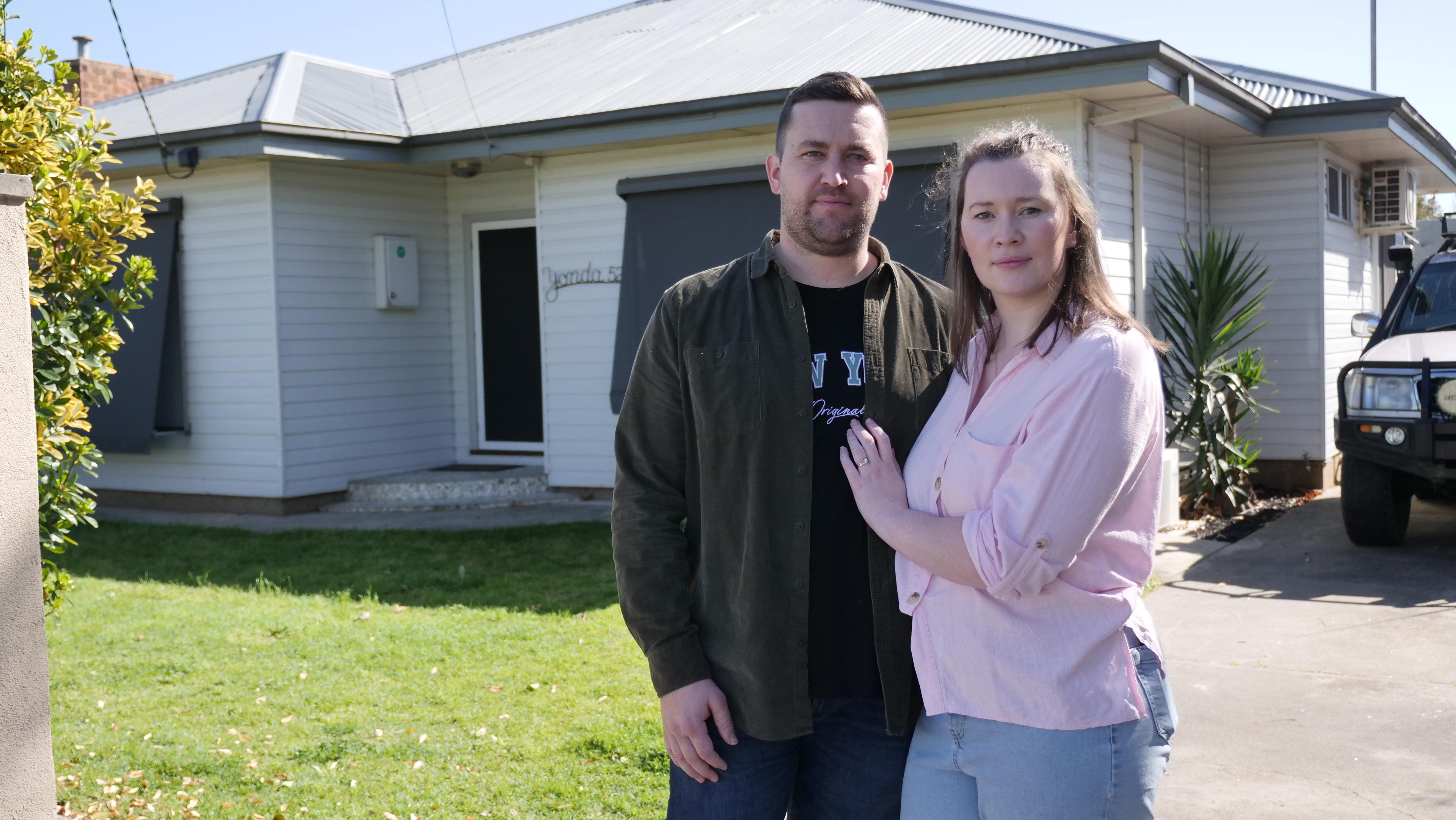 Megan Raines stands next to her partner Brandon with her hand on his chest both looking serious in front of their home