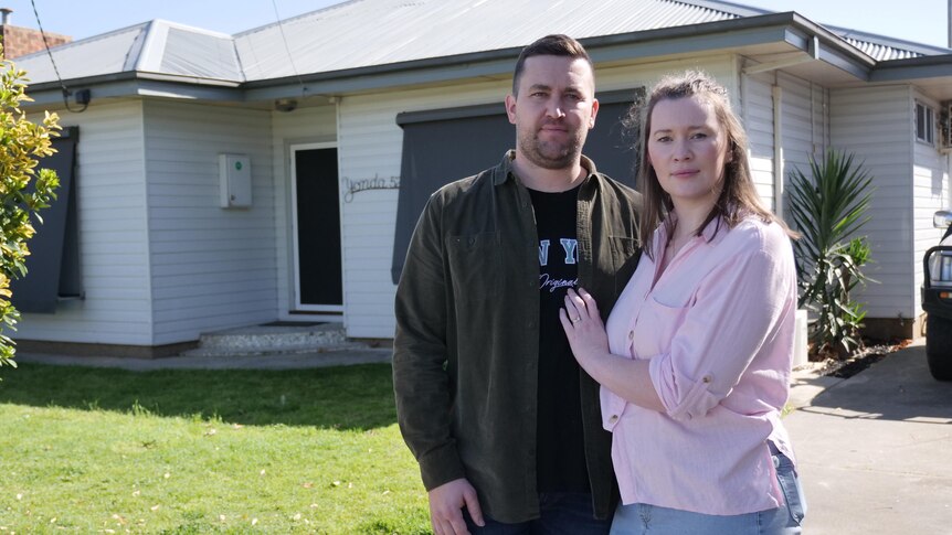 Megan Raines stands next to her partner Brandon with her hand on his chest both looking serious in front of their home