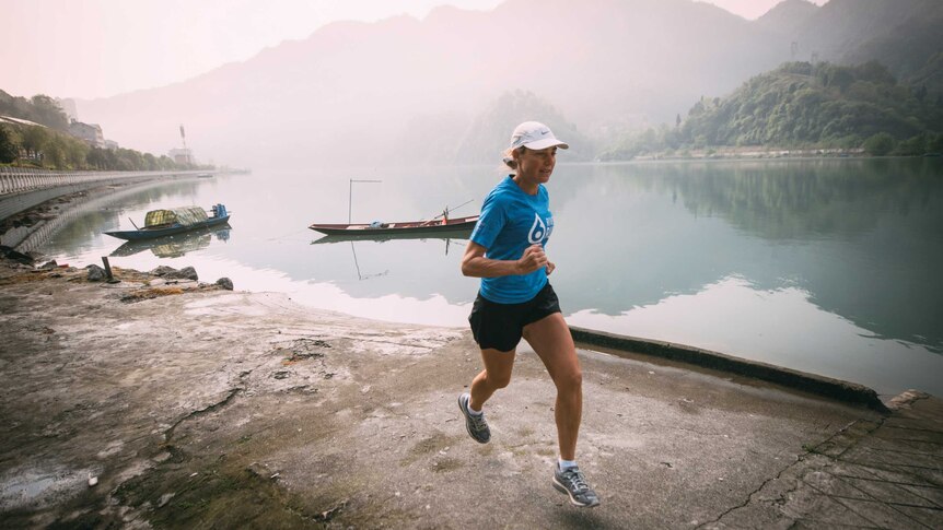 Woman runs along river with thick haze and mountain in the background.