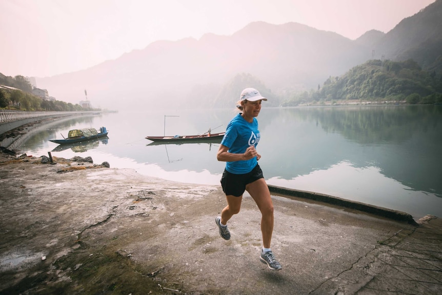 Woman runs along river with thick haze and mountain in the background.