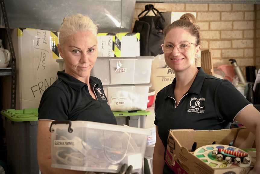 Two women in black polo shirts look at the camera while carrying boxes.