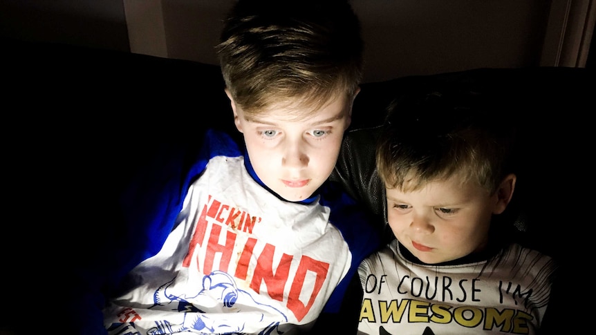 Two young boys sit on a couch, their faces lit up by an electronic device