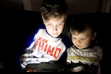 Two young boys sit on a couch, their faces lit up by an electronic device