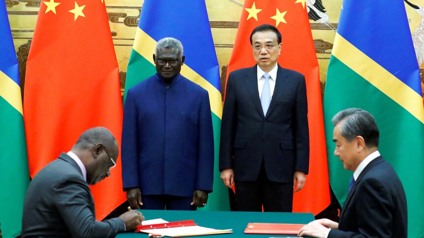 Manasseh Sogavare and Li Keqiang smile in front of Solomon Islands and Chinese flags as people sign documents.