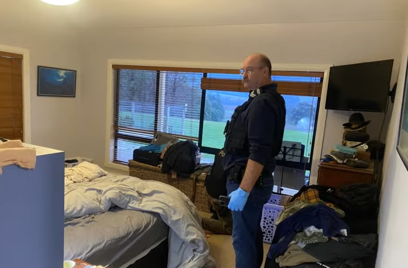 A police officer in a navy uniform with blue plastic gloves on his hands stands side-on in a bedroom of a house.
