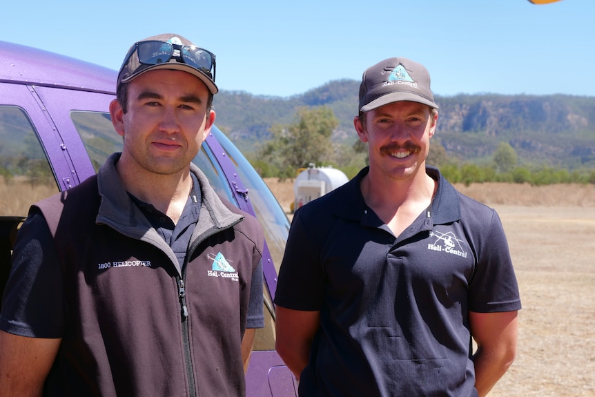 Patrick and Eiran standing in front of a purple helicopter looking at the camera, ranges, grass and trees behind.