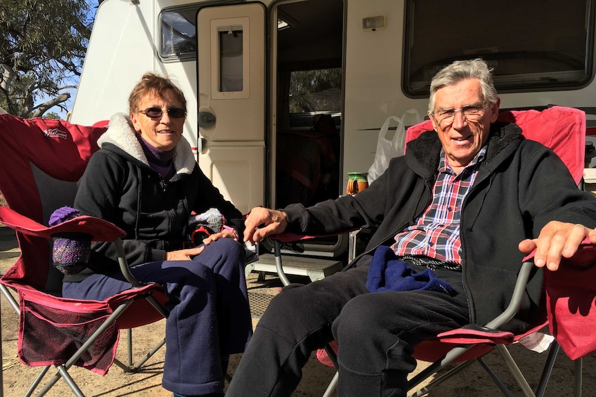 Two retirees sit in fold-out chairs in front of a caravan
