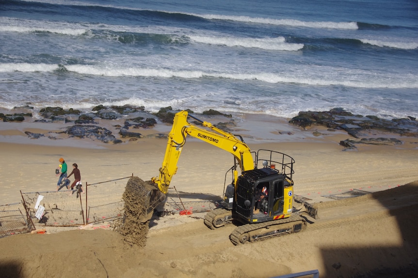 Deux personnes marchent le long d'une plage près d'une machine à creuser qui ramasse du sable.