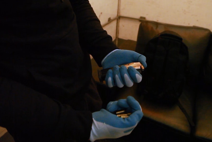 Gloved hands hold a gun's ammunition clip and a handful of bullets.