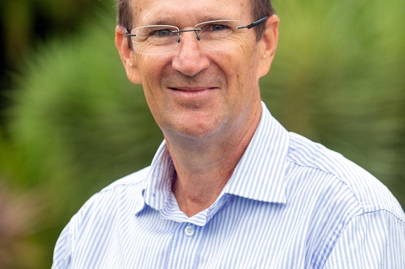 A man in glasses and a collared shirt sits in front of a green vegetation background. 