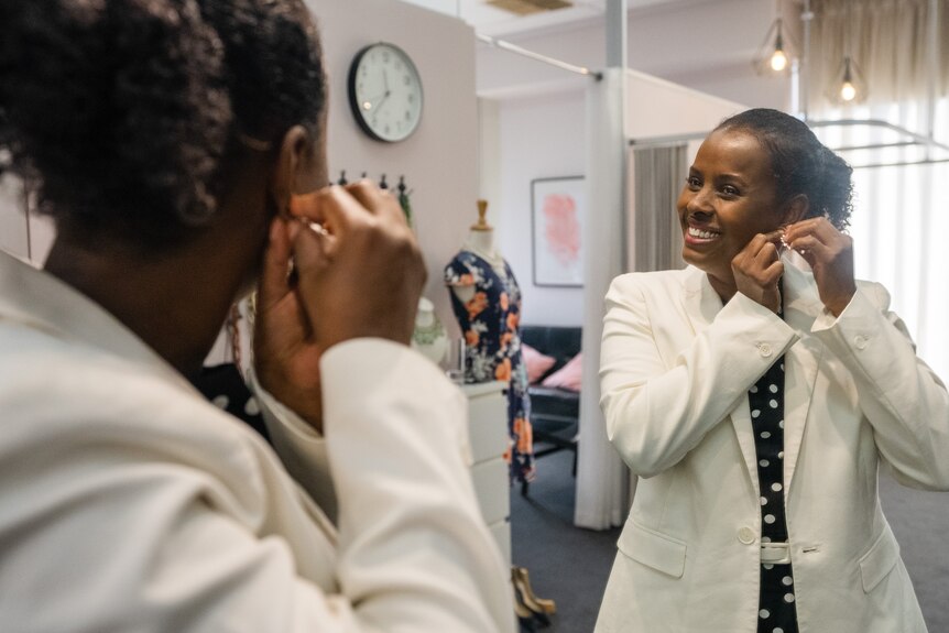 A woman wearing a white suit jacket is putting on an earing, smiling into a full length mirror.