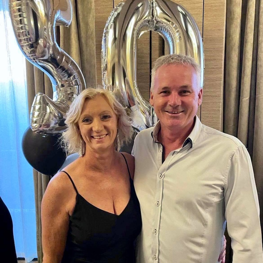 A woman with blonde hair and wearing a black top stands smiling with a man with short white hair wearing a collared shirt.