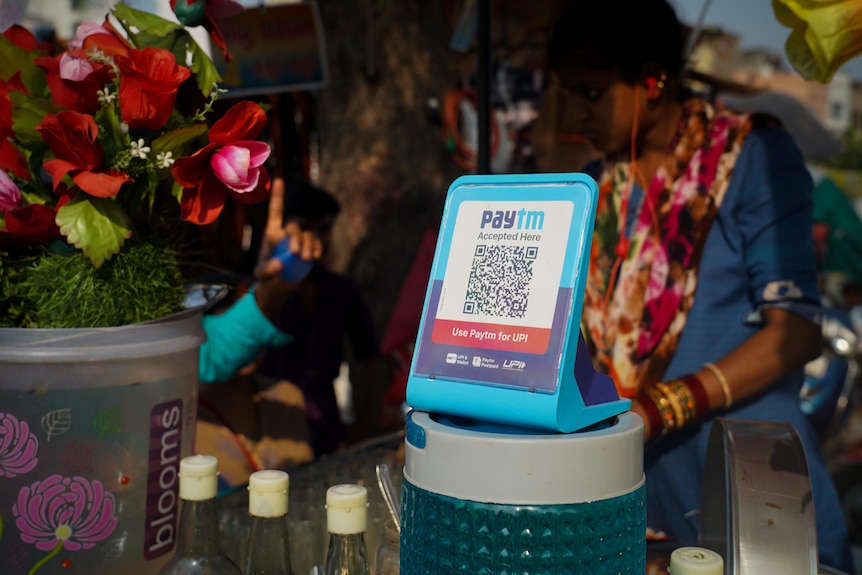 A QR code sign sits atop a jar next to a bunch of flowers at a market stall
