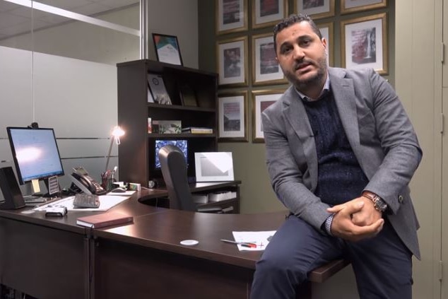 A man sitting on a desk talking to camera.