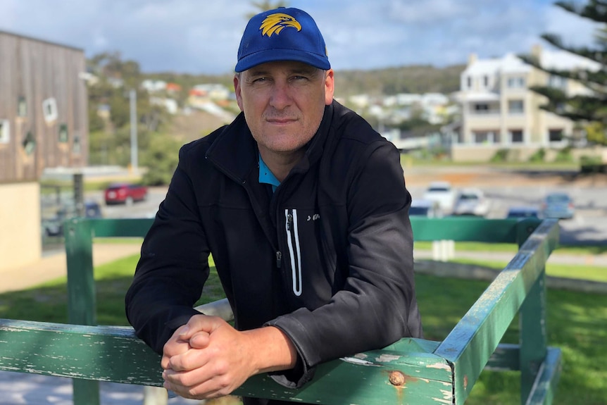 A man in a black jacket and West Coast Eagles cap leans on a green wooden fence.