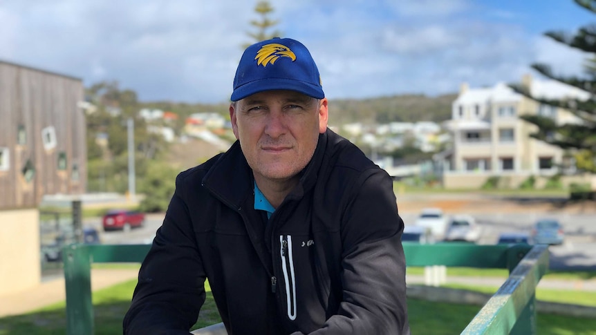 A man in a black jacket and West Coast Eagles cap leans on a green wooden fence.