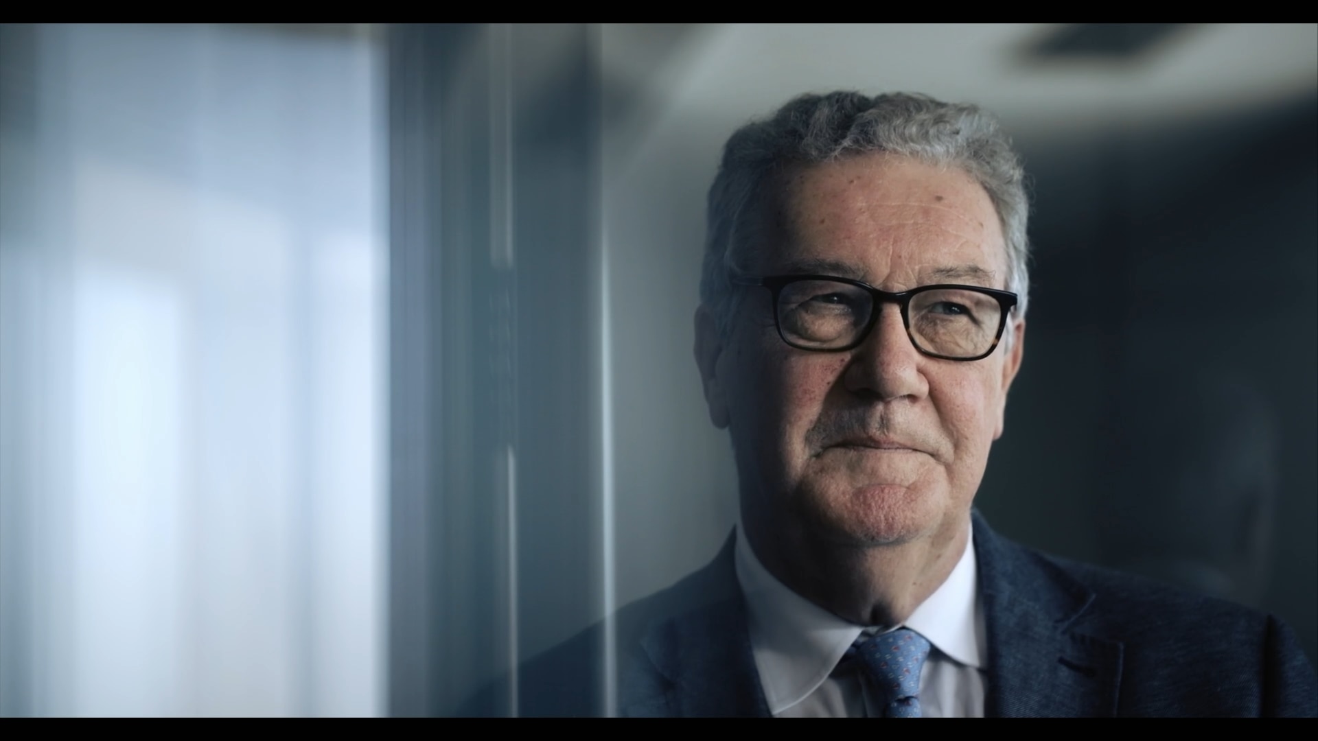 A medium close-up shot of an older man with grey hair, glasses and a wry expression.