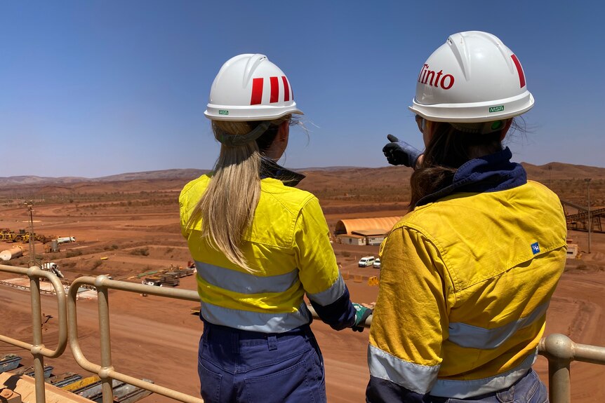 Dos mujeres con camisas de color amarillo brillante y cascos blancos miran por encima de una mina de mineral de hierro.