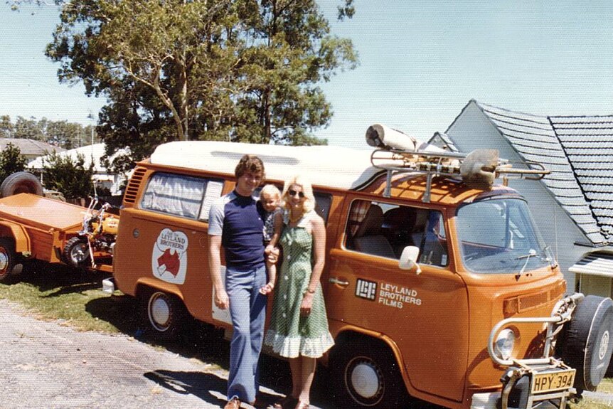 An old photo of a man, woman and baby in front of an orange VW Kombi van, blue sky, tree.