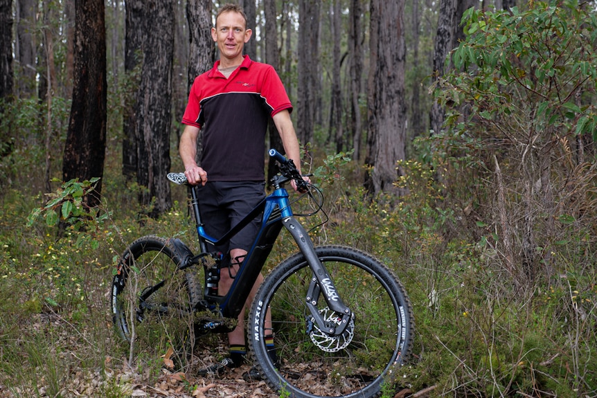 A man leaning on a blue mountain bike surrounded by trees and shrubs