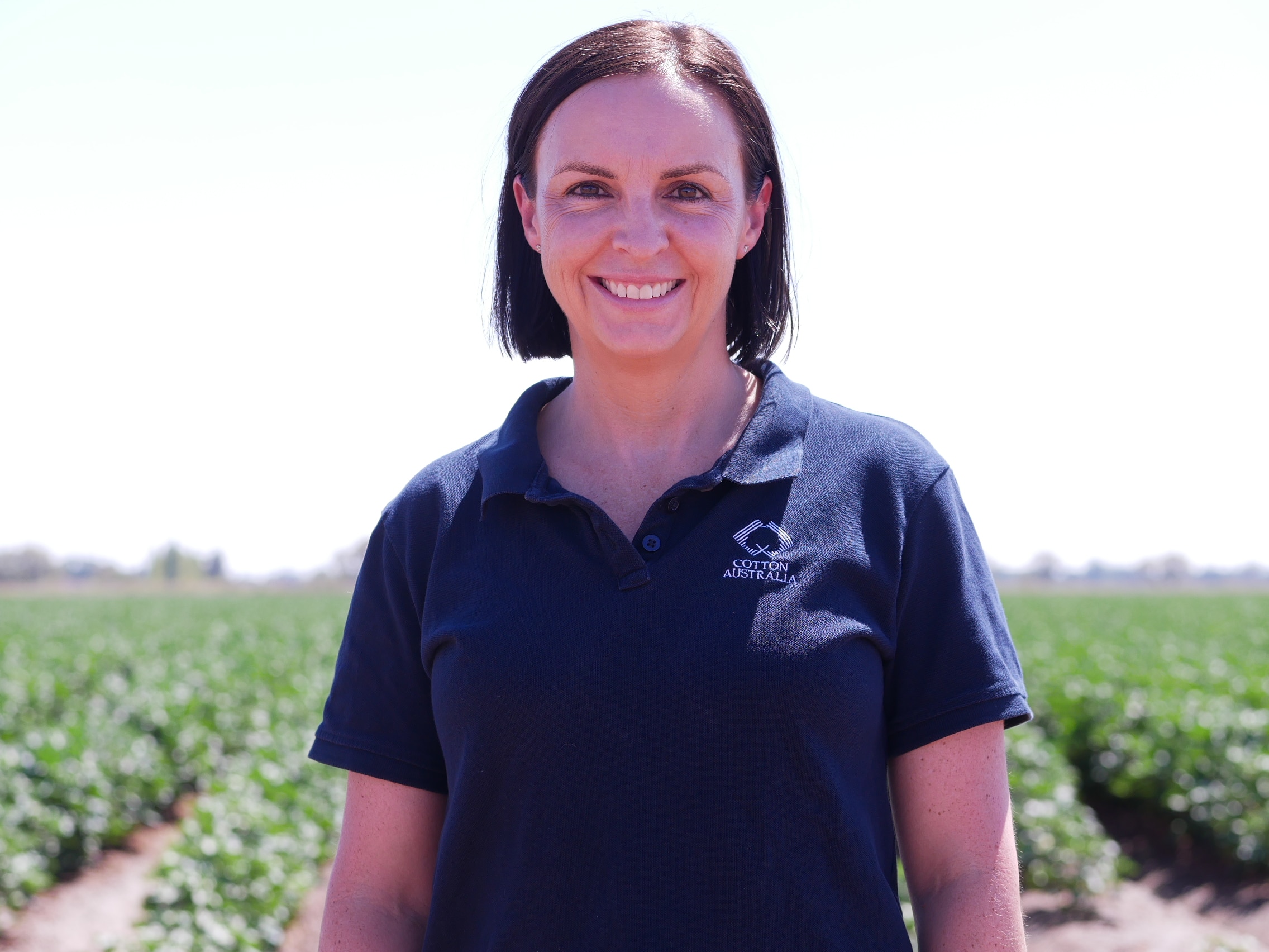 Una mujer de cabello castaño sonríe a la cámara; al fondo se pueden ver hileras de algodón.