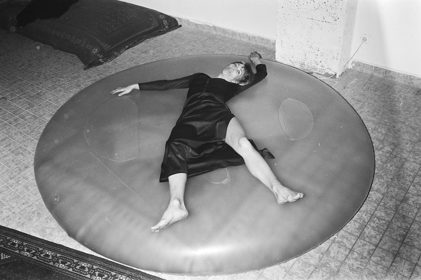 Black and white photo, a woman lies on a round vinyl waterbed wearing a dark dress