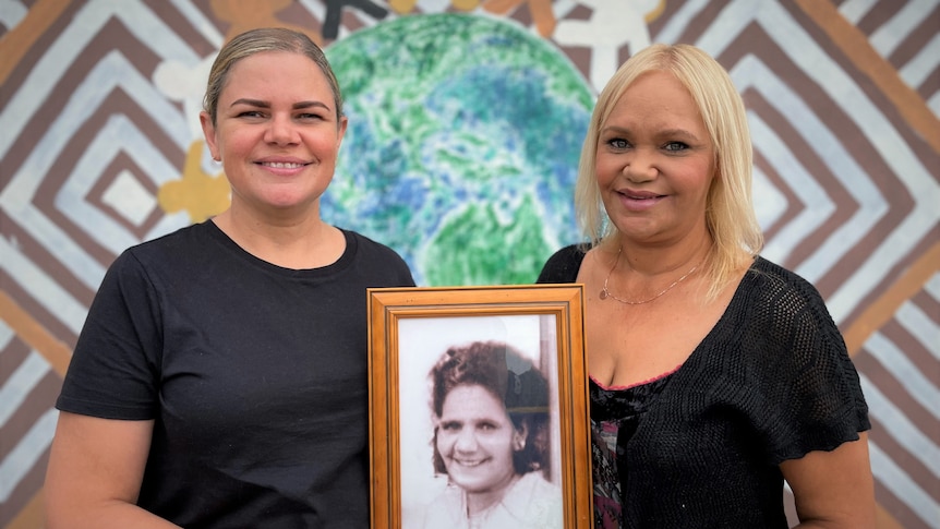 Two women stand in front of Aboriginal artwork, holding an old framed photo of a woman