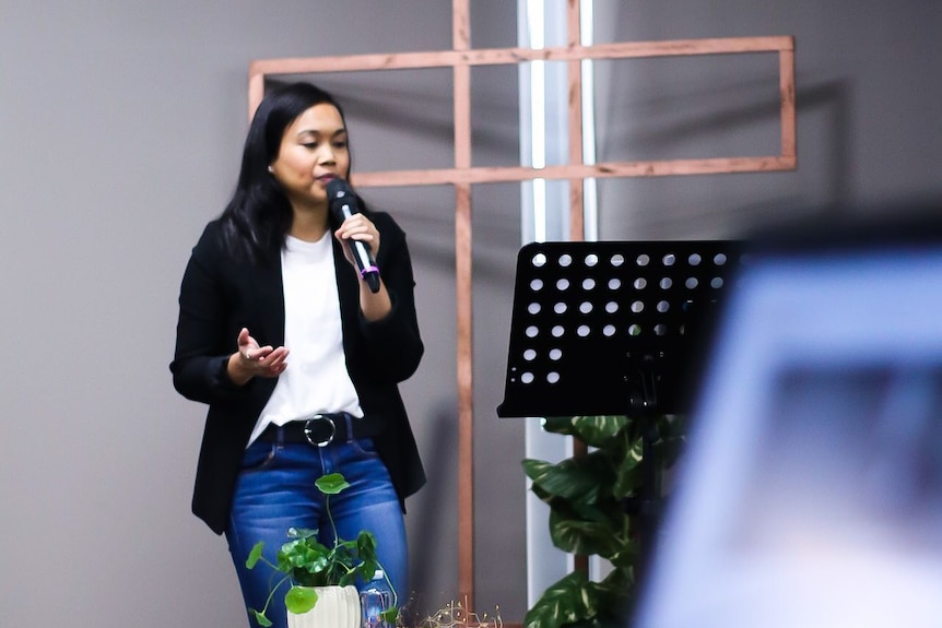 A young woman speaks into a microphone in front of a wooden crucifix.