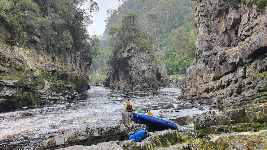 a man in high vis wet weather gear with a back pack sits next to his raft on a rock facing a rocky outcrop at a bend in a river