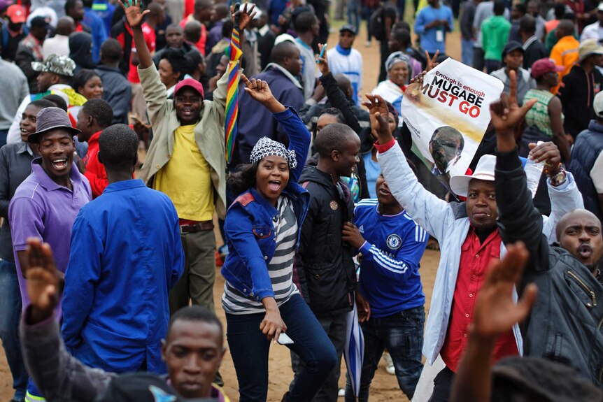 A group of colourfully dressed people walk through the streets of Harare, one holds a poster saying "Mugabe must go".