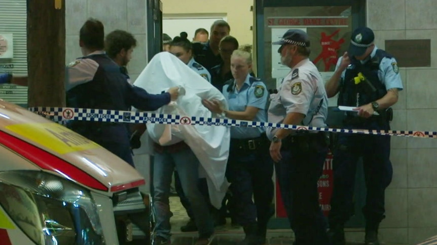 A man under a white blanket being led away by police