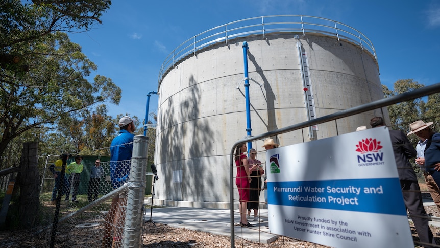 A water tank inside a fence with a sign for NSW Water, with people standing in front of it.