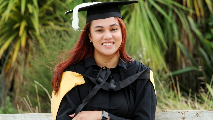 A woman in a graduation cap and gown sits smiling in front of green palm trees 