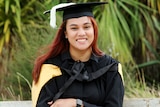 A woman in a graduation cap and gown sits smiling in front of green palm trees 