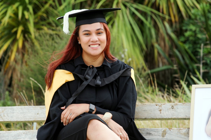 A woman in a graduation cap and gown sits smiling in front of green palm trees 